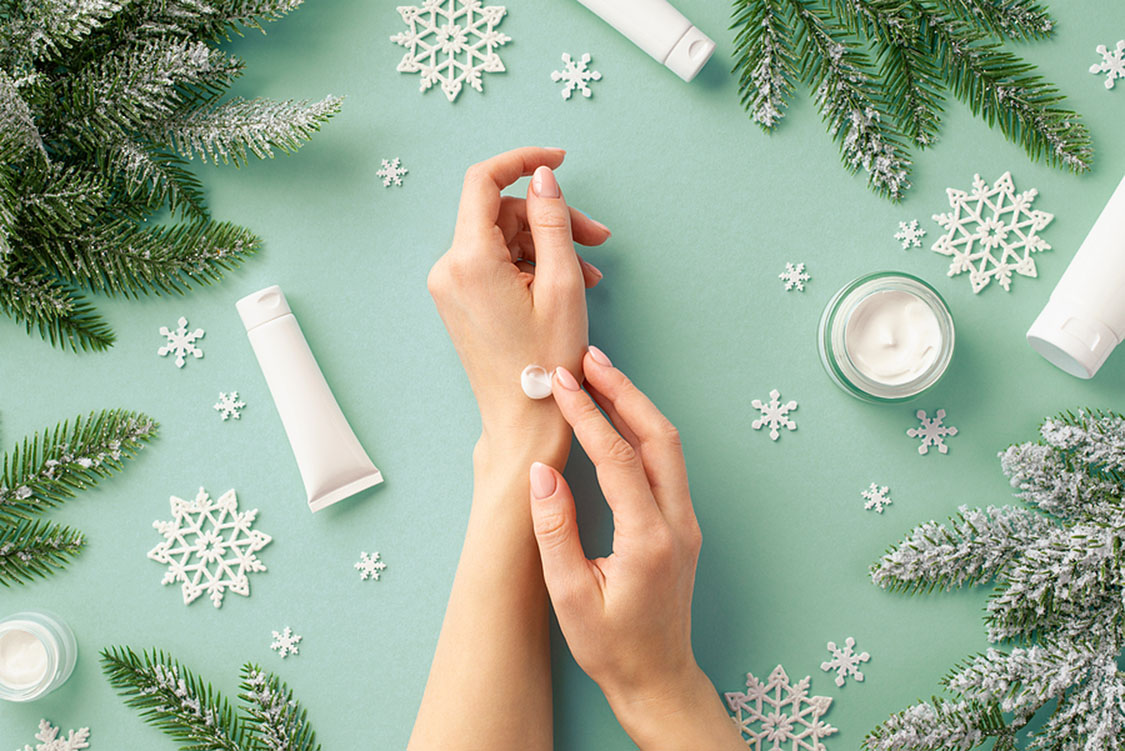 Winter season skin care concept. First person top view photo of woman using hand cream cosmetic jars tubes snowflakes and spruce branches in hoarfrost on isolated pastel green background; winter skin care tips